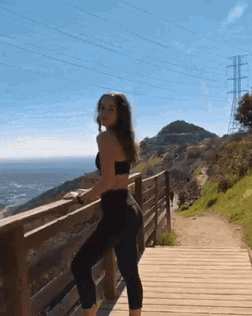 a woman in a black top and black leggings is standing on a wooden deck overlooking the ocean .