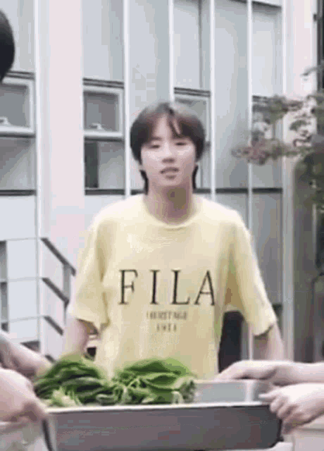 a young boy wearing a yellow fila t-shirt is standing in front of a tray of greens .
