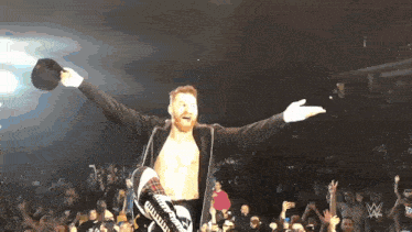 a wrestler with his arms outstretched stands in front of a crowd with a w logo in the background