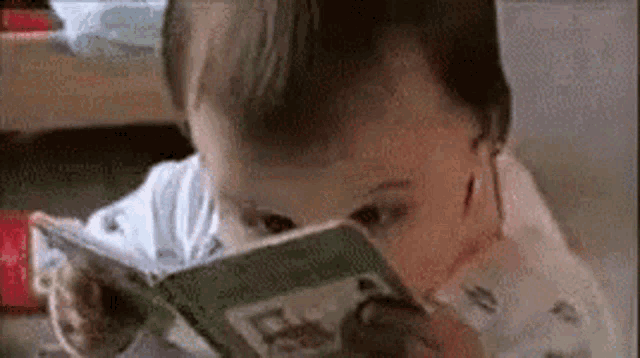 a baby is reading a book with his eyes closed while sitting at a table .