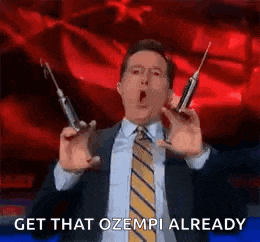 a man in a suit and tie is holding two syringes and says `` get that ozempi already '' .