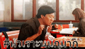 a man is sitting at a table eating noodles with a straw while a woman sits behind him