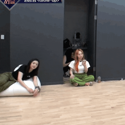 two women are sitting on the floor in front of a sign that says glow up