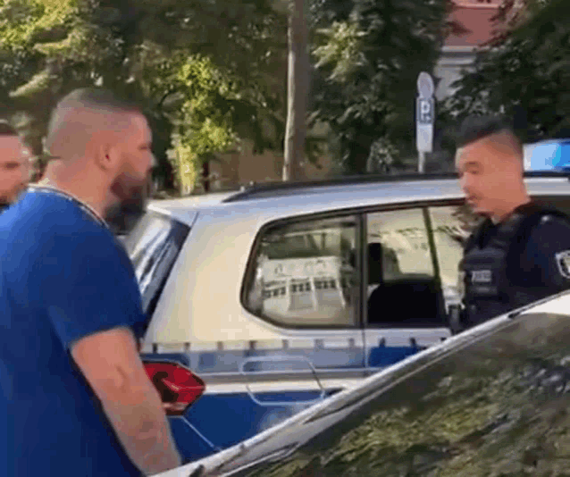 a man in a blue shirt is standing in front of a police car talking to a police officer .