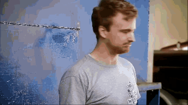 a man with a beard wearing a grey t-shirt with the word army on it stands in front of a blue wall