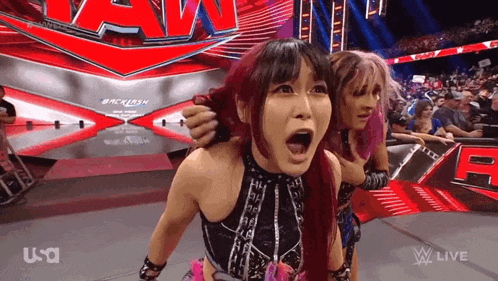 two female wrestlers are standing on a stage with their mouths open in front of a crowd at a wrestling event .