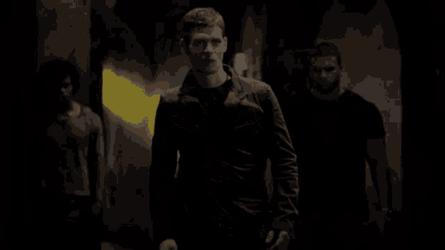 a man in a black jacket is walking down a dark hallway with two other men .