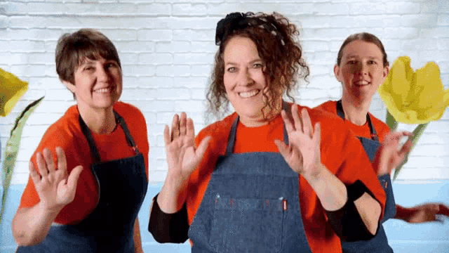 three women wearing aprons and orange shirts are dancing in front of a white brick wall