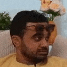 a man wearing sunglasses is sitting on a couch with a vase of flowers in the background .