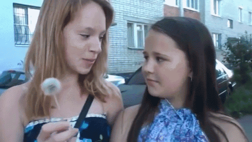 two young girls are looking at each other while one girl blows a dandelion
