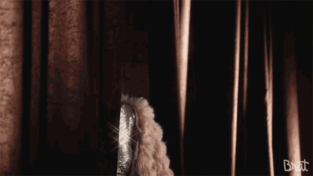 a woman in a fur coat is peeking out from behind a curtain that says brat on the bottom