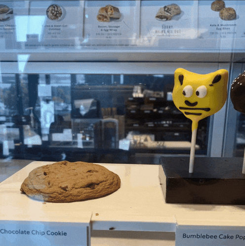 a chocolate chip cookie and a bumblebee cake pop are on display in a bakery