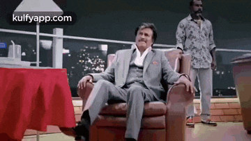 a man in a suit is sitting in a chair while another man stands in the background .