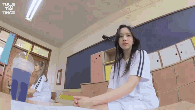 a girl is sitting at a desk in a classroom with a cup of drink .