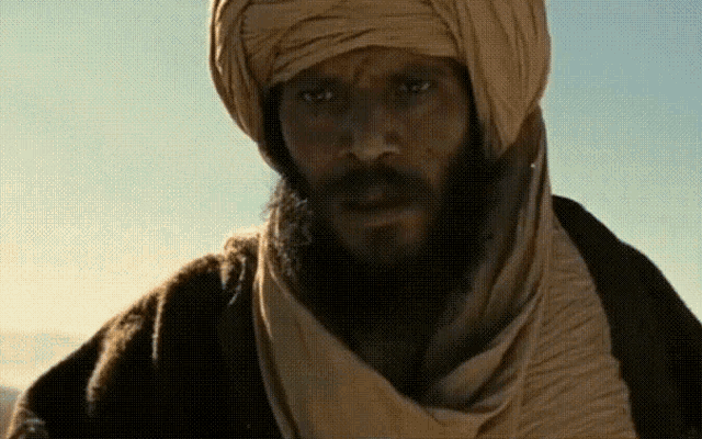 a man with a beard wearing a turban and scarf looks at the camera