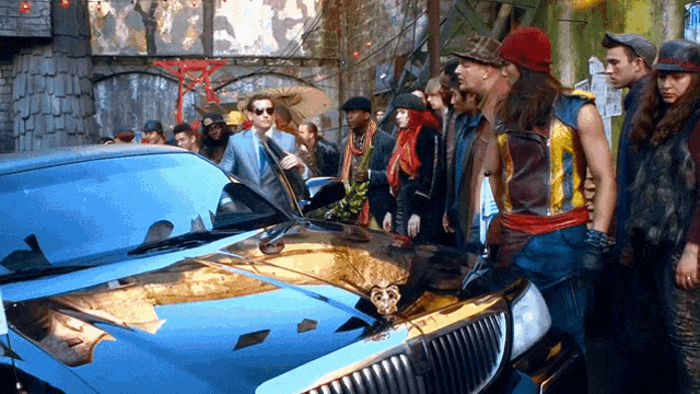 a group of people are standing around a blue car with a gold emblem on the hood