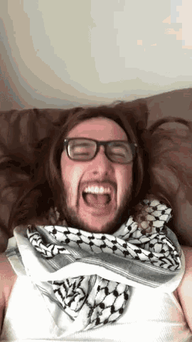 a man with glasses and a scarf around his neck is laying on a bed with his mouth open .