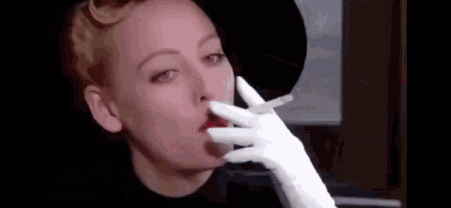 a woman is smoking a cigarette in a black hat and white gloves .
