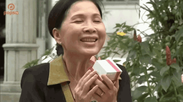 a woman is smiling while holding a small white box with a red ribbon in front of a sign that says popsa