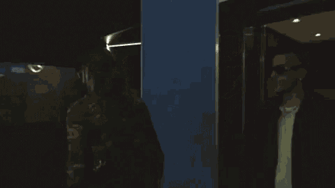 a man wearing sunglasses and a black jacket is standing in a dark room in front of a mirror .