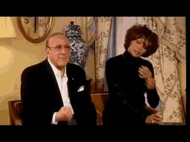 a man in a tuxedo and a woman in a black turtleneck are standing next to each other in a living room