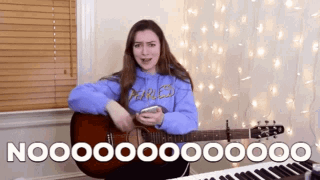 a woman in a blue hoodie is playing a guitar and singing while holding a cell phone