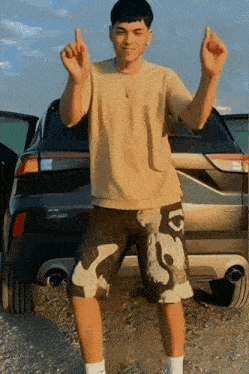 a young man in a tan shirt and camo shorts stands in front of a black suv