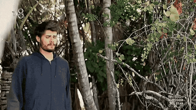 a man in a blue hoodie is standing in a forest with a tv8 logo above him