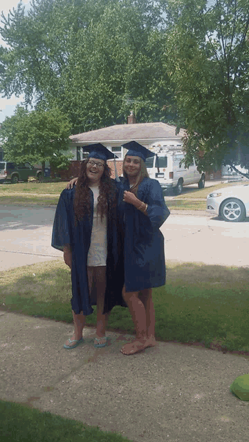 two girls in graduation gowns pose for a picture in front of a house
