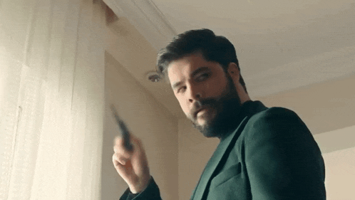 a man with a beard in a green suit is standing in front of a window holding a knife .