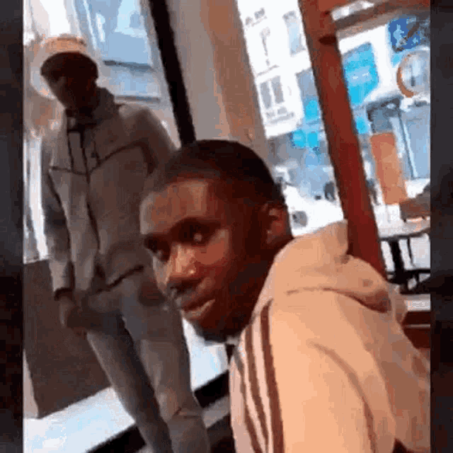 a man in a white hoodie is sitting in front of a window with another man standing behind him .