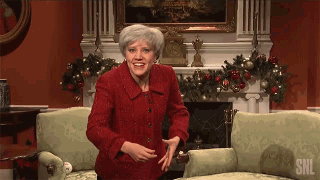 a woman in a red jacket is dancing in front of a fireplace with the snl logo on the couch
