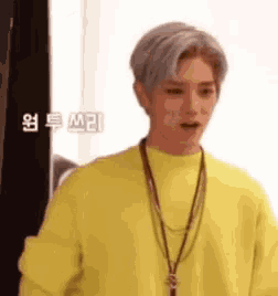 a man wearing a yellow sweater and a necklace is standing in front of a black curtain .