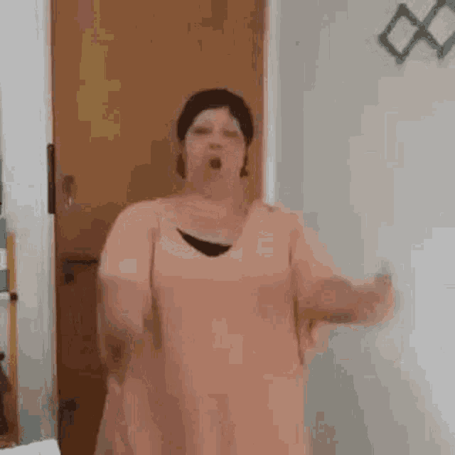 a woman in a pink dress is standing in front of a door and making a funny face .