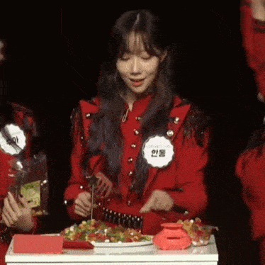 a woman in a red jacket is standing in front of a table with a plate of food on it .