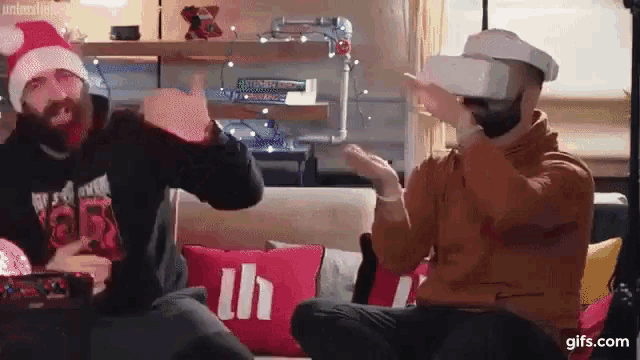 two men wearing santa hats and virtual reality headsets are sitting on a couch in a living room .