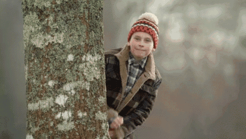 a young boy is peeking out from behind a tree wearing a hat and jacket .