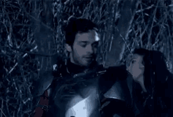 a man in armor is standing next to another man in a dark forest .