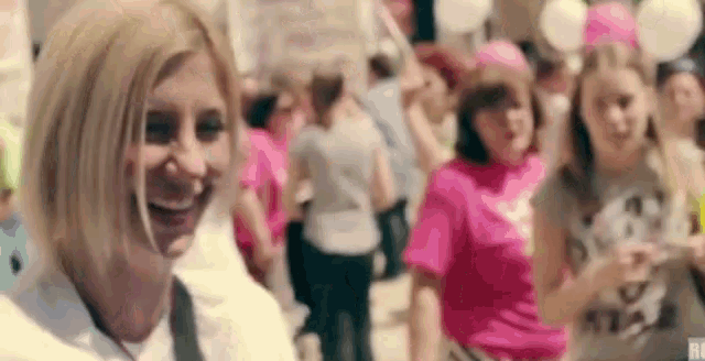 a woman is smiling in front of a crowd of people wearing pink shirts and balloons .