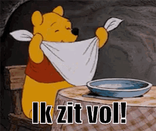 winnie the pooh is sitting at a table with a towel around his neck and the words " ik zit vol " below him .