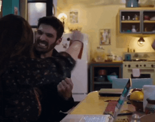 a man and a woman are hugging in a kitchen with a laptop on the table .
