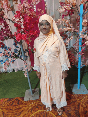a woman wearing a hijab and glasses stands in front of a tree with pink flowers