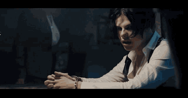 a man in a white shirt and suspenders is sitting at a table with his hands folded in front of him .