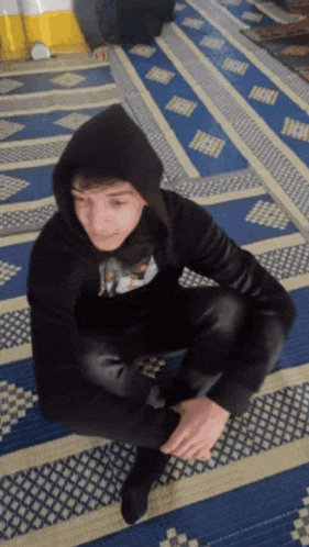 a man in a black hoodie sits on a blue rug