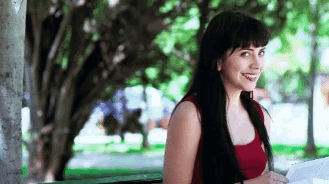 a woman in a red tank top smiles while sitting on a park bench