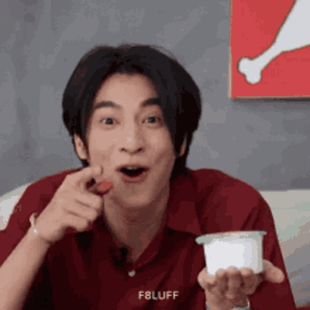a young man in a red shirt is eating a strawberry while holding a container of yogurt .