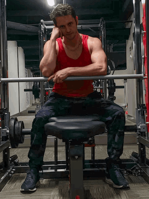a man in a red tank top sits on a bench holding a barbell in a gym