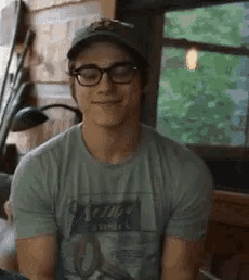 a young man wearing glasses and a hat is smiling while sitting in front of a window .