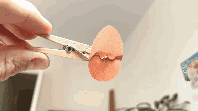 a person is holding a paper egg in a pair of wooden clothespins