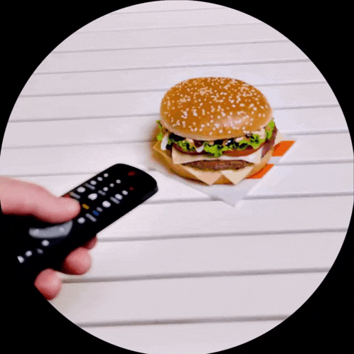a person is holding a remote control in front of a hamburger on a table